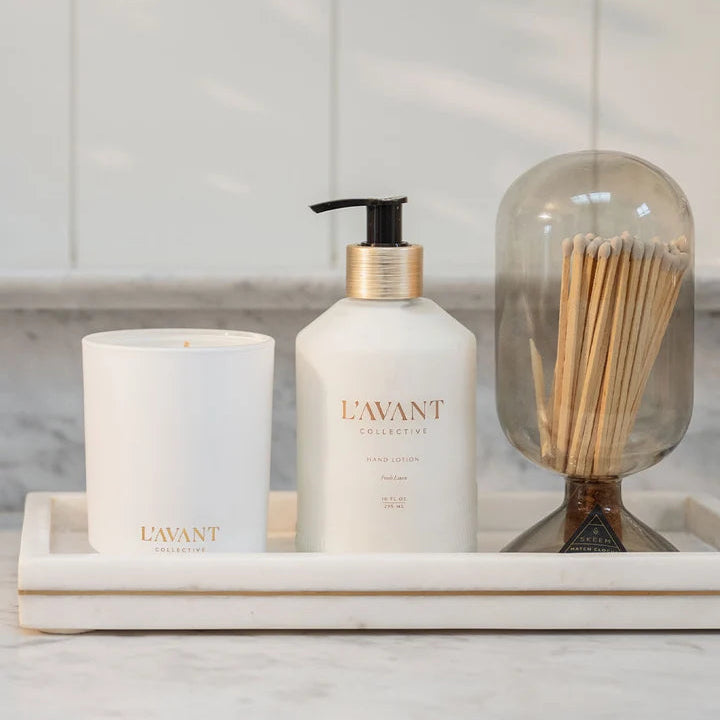 L’AVANT Collective Ambre Santal candle styled with hand lotion and decorative matches on a marble vanity tray.