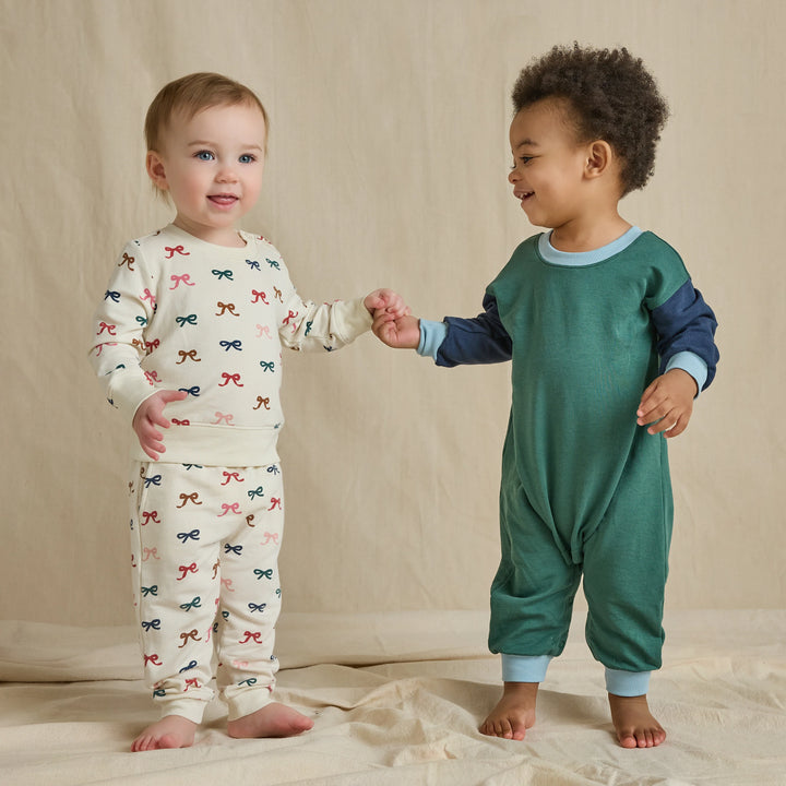 Baby wearing a cream multicolor bow sweatshirt and jogger set alongside another child in a green outfit