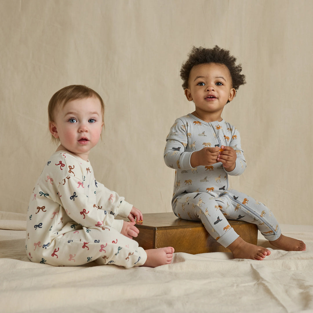 Baby wearing a cream multicolor bow romper while sitting beside another baby in a printed one-piece outfit