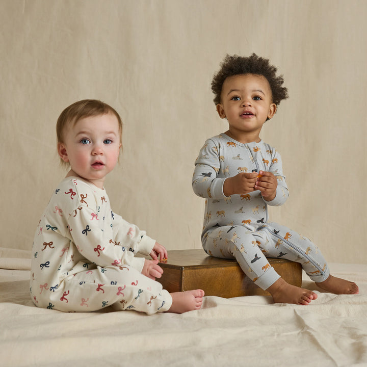 Baby wearing a cream multicolor bow romper while sitting beside another baby in a printed one-piece outfit