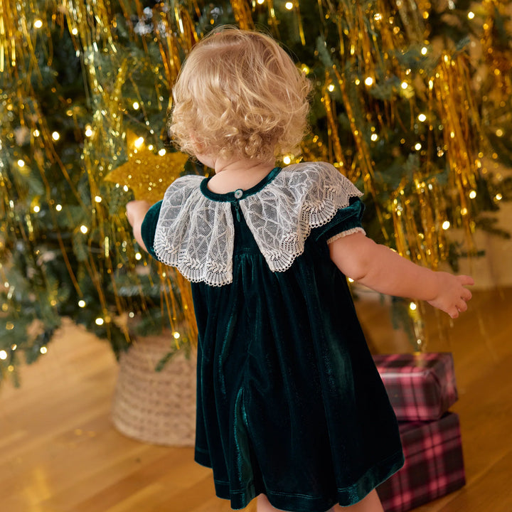 Back view of baby girl wearing the Evergreen Velour Caroline Dress with a white lace collar beside a festive Christmas tree