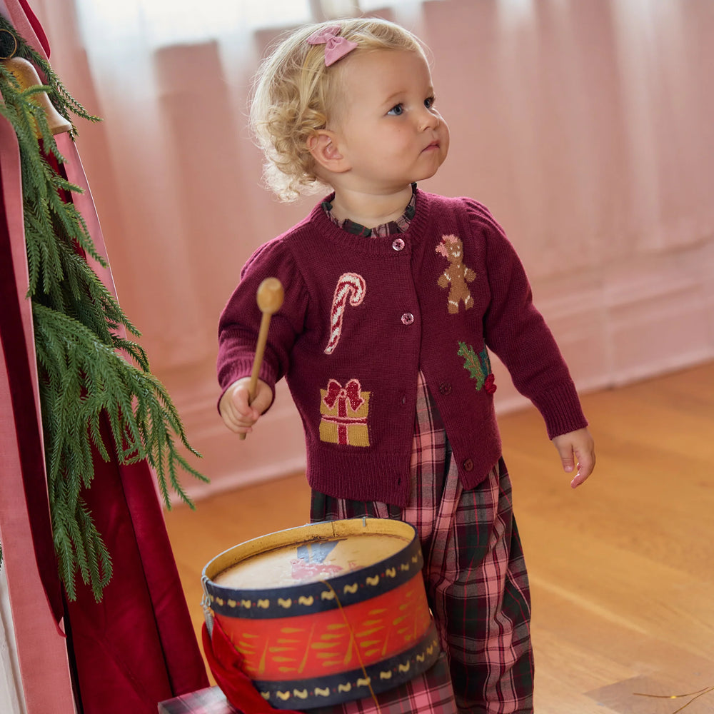 Baby girl wearing the Pink Chicken Constance Sweater in burgundy with a festive plaid outfit while playing with a Christmas drum.