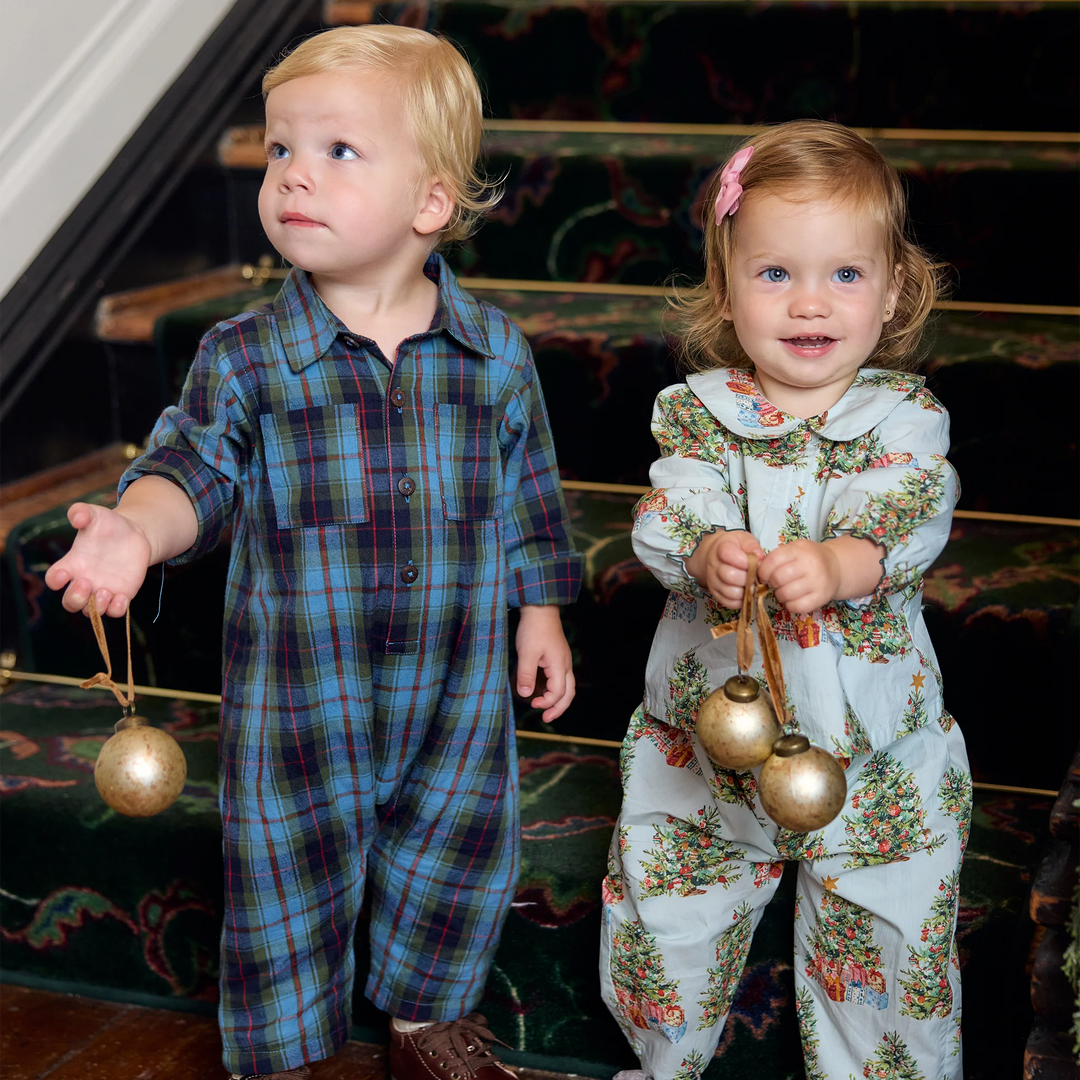 Baby girl wearing the Pink Chicken Dawson 2PC Set in Trees and Presents alongside a toddler in blue plaid, holding gold Christmas ornaments.