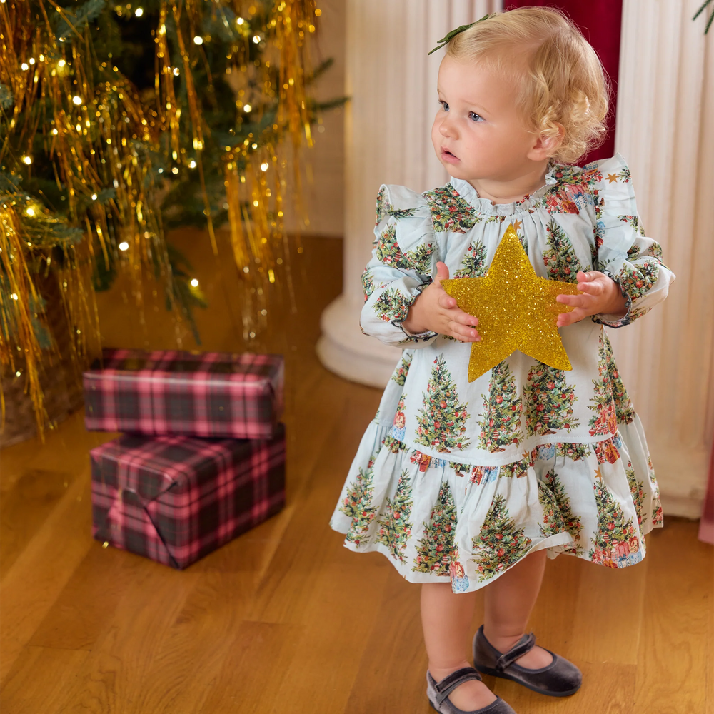 Baby girl wearing the Pink Chicken Harper Dress Set in Trees and Presents while holding a gold glitter star beside a decorated Christmas tree.