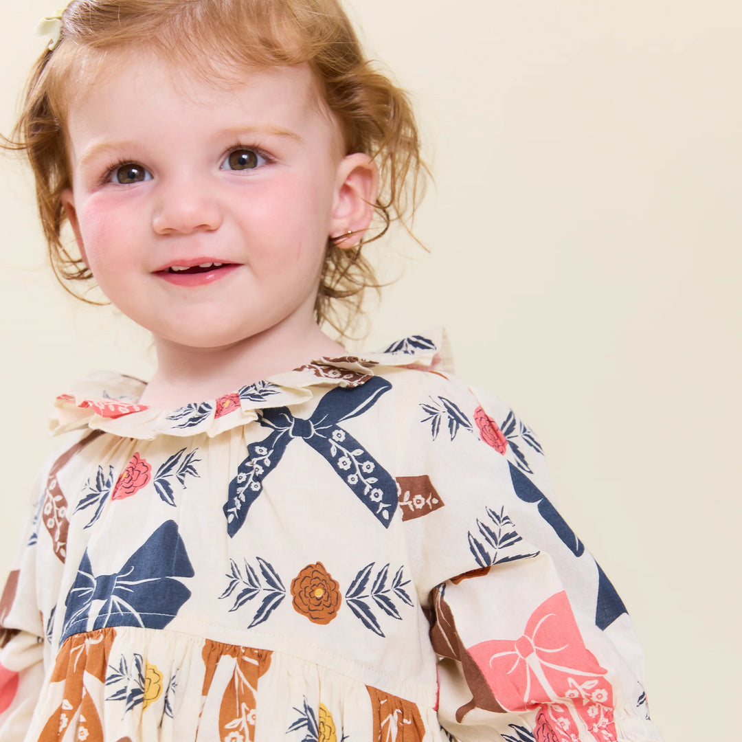 Baby girl wearing cream Harvest Bow Dress with ruffled neckline, floral accents and colorful bows