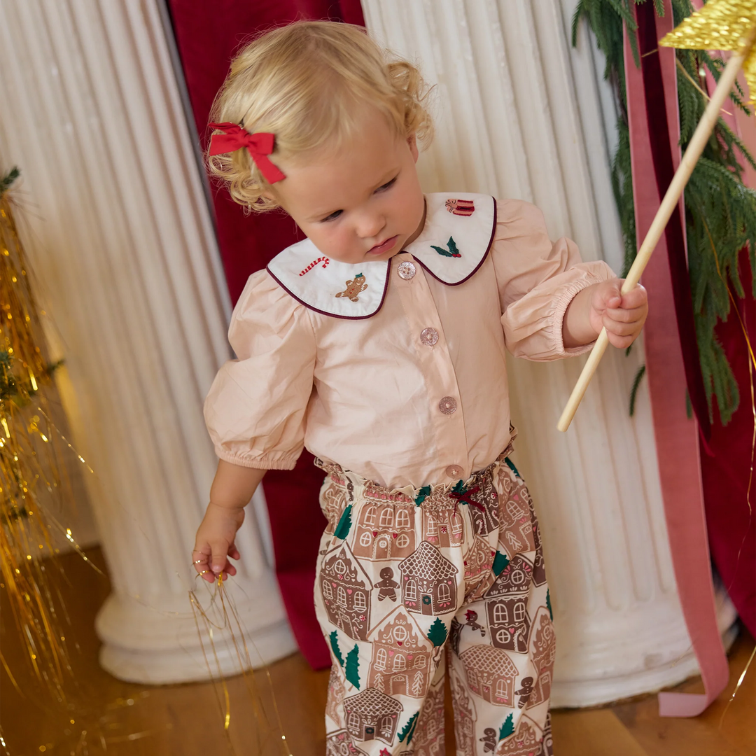 Little girl modeling the Pink Chicken Noelle 2PC Set with a pink embroidered collar top, gingerbread print pants, and red hair bow.