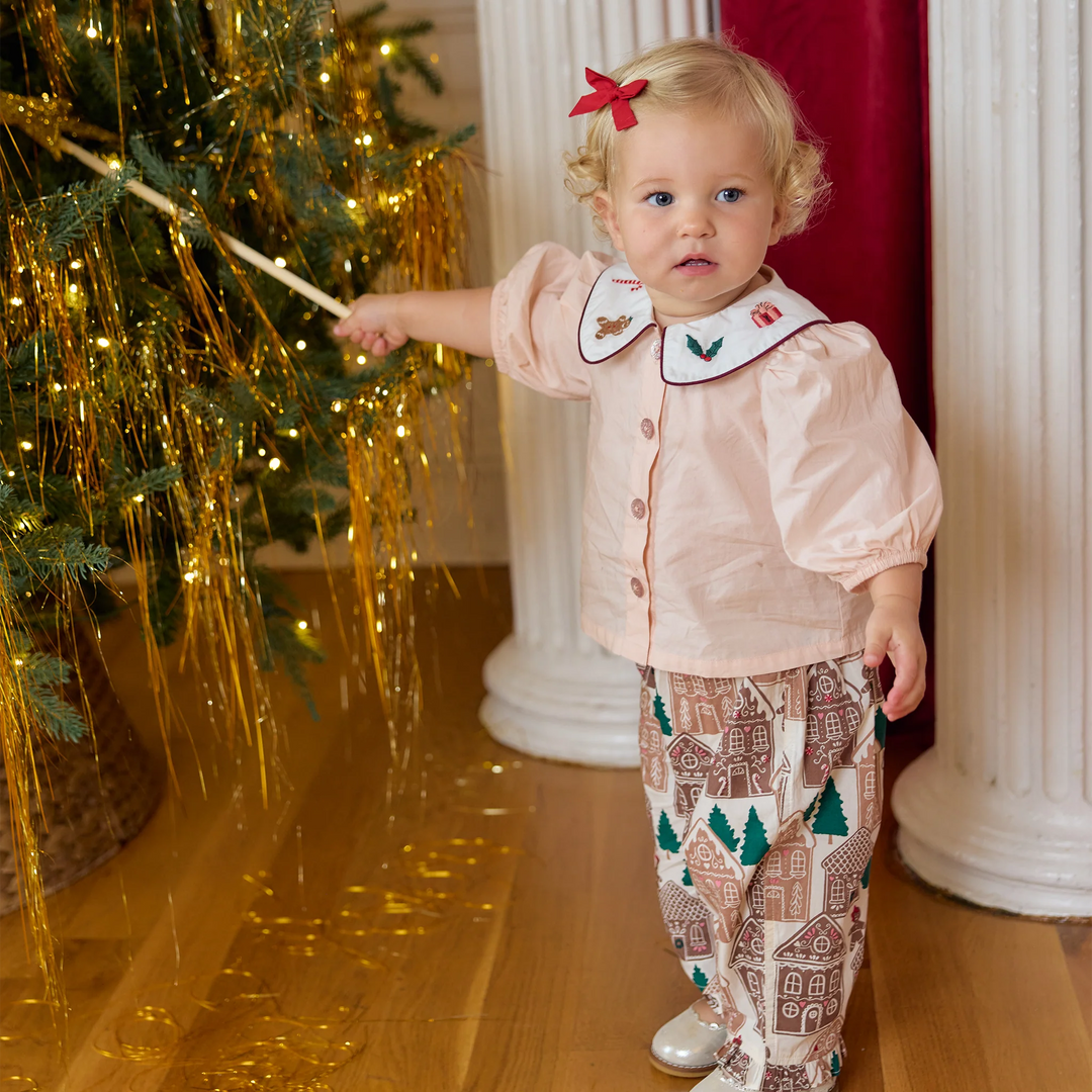 Toddler girl wearing the Pink Chicken Noelle 2PC Set in Gingerbread Houses while standing beside a decorated Christmas tree.