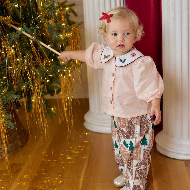 Toddler girl wearing the Pink Chicken Noelle 2PC Set in Gingerbread Houses while standing beside a decorated Christmas tree.