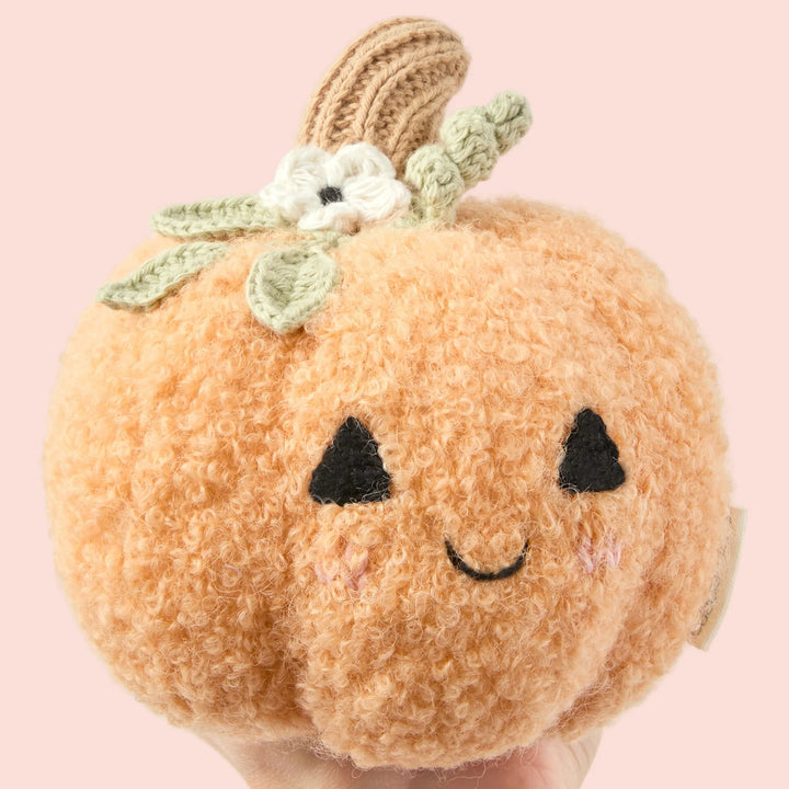 Baby Pumpkin Moonflower orange plush toy with smiling embroidered face and floral knit details on a pink background