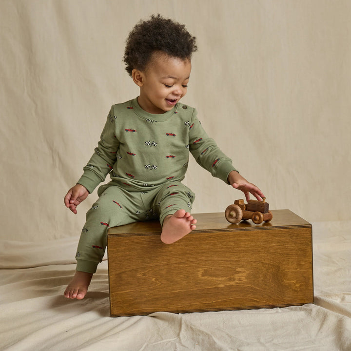 Toddler boy wearing an olive green race car and checkered flag sweatshirt and jogger pants set while playing with a wooden toy car.
