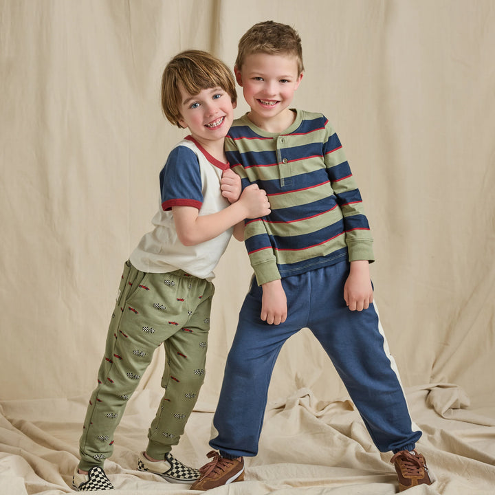 Boy wearing an olive green, navy, and red striped long sleeve Henley shirt styled with blue jogger pants.