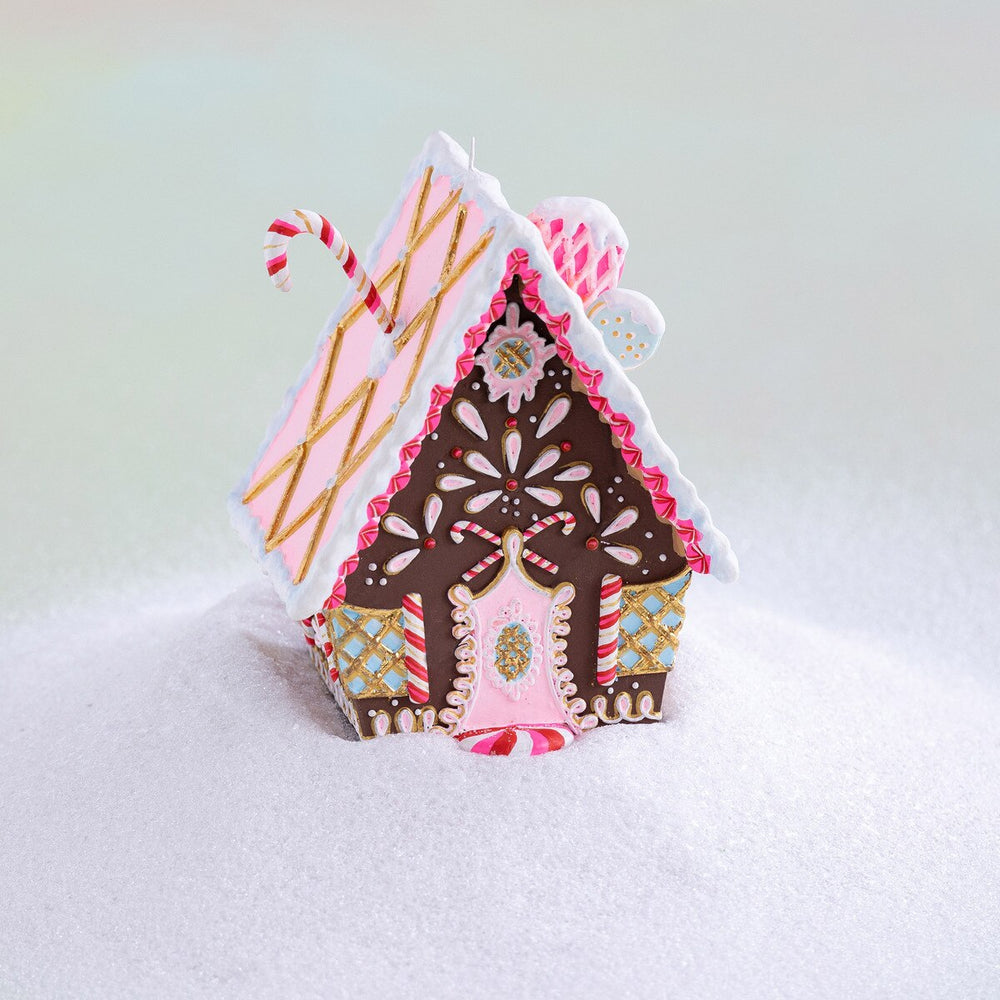 Brown gingerbread cottage ornament with pink and white icing, candy cane accents, gold details, and a pink door, displayed on a snowy background.