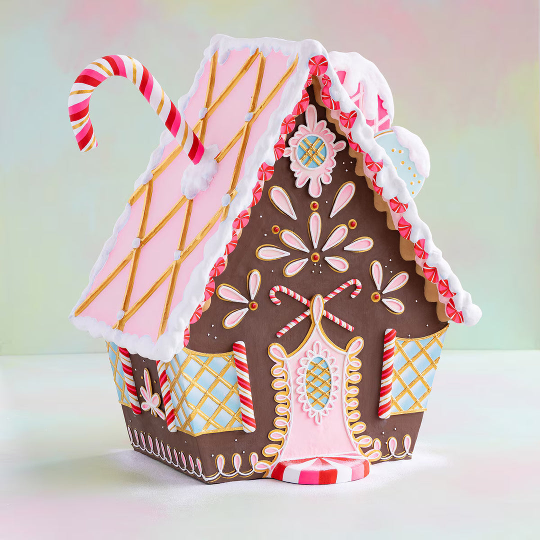 Brown gingerbread cottage with pink and white icing, candy cane accents, gold details, and a pink door.