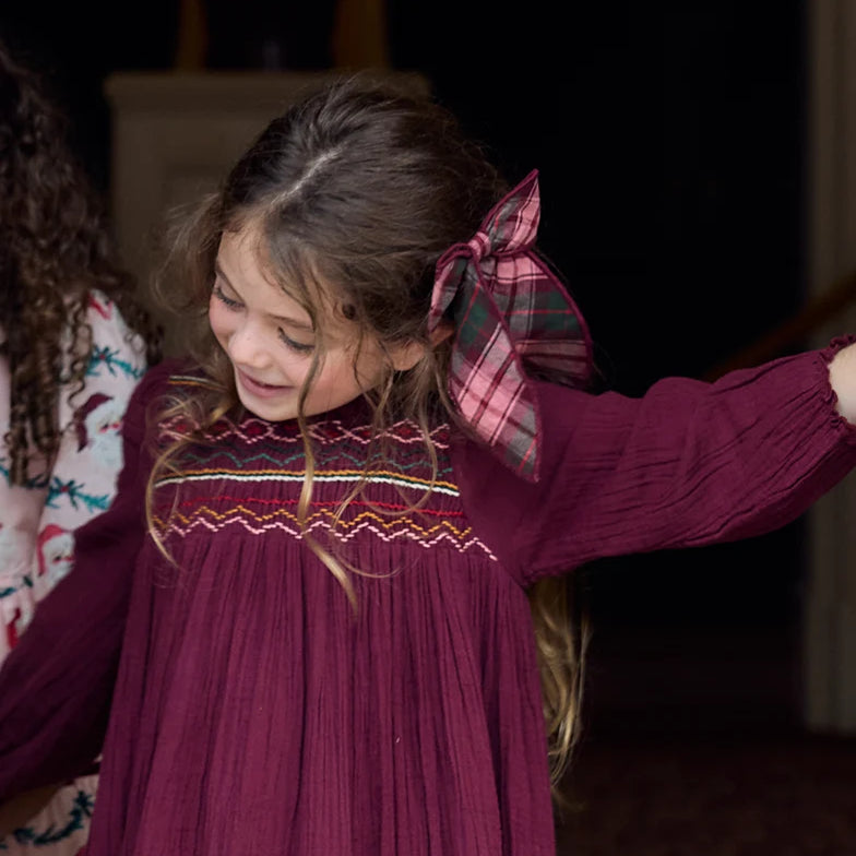 Young girl wearing the Pink Chicken Burgundy Tartan Long Tail Bow in her hair, styled with a burgundy smocked dress for a festive holiday look.