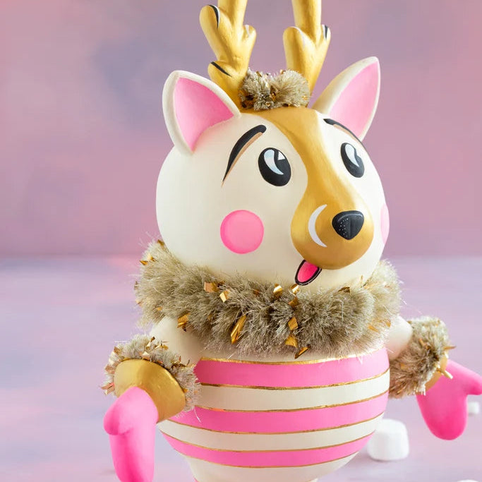 Close-up of a cartoon-like La Renne reindeer with cream-white fur, pink cheeks and ears, gold antlers, a gold-accented face, fluffy gold tinsel collar, and pink-and-gold striped body against a soft pink backdrop.