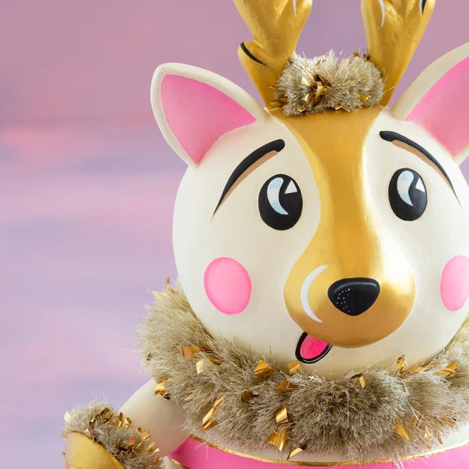 Close-up of a cartoon-like La Renne reindeer with a white and gold face, pink cheeks and ears, large expressive eyes, gold antlers, and fluffy gold tinsel trim against a soft pink backdrop.