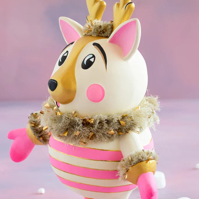 Side profile of a cartoon-like La Renne reindeer with cream-white fur, pink inner ears and cheeks, gold antlers and muzzle, fluffy gold tinsel collar, and a pink-and-gold striped body against a soft pink backdrop.