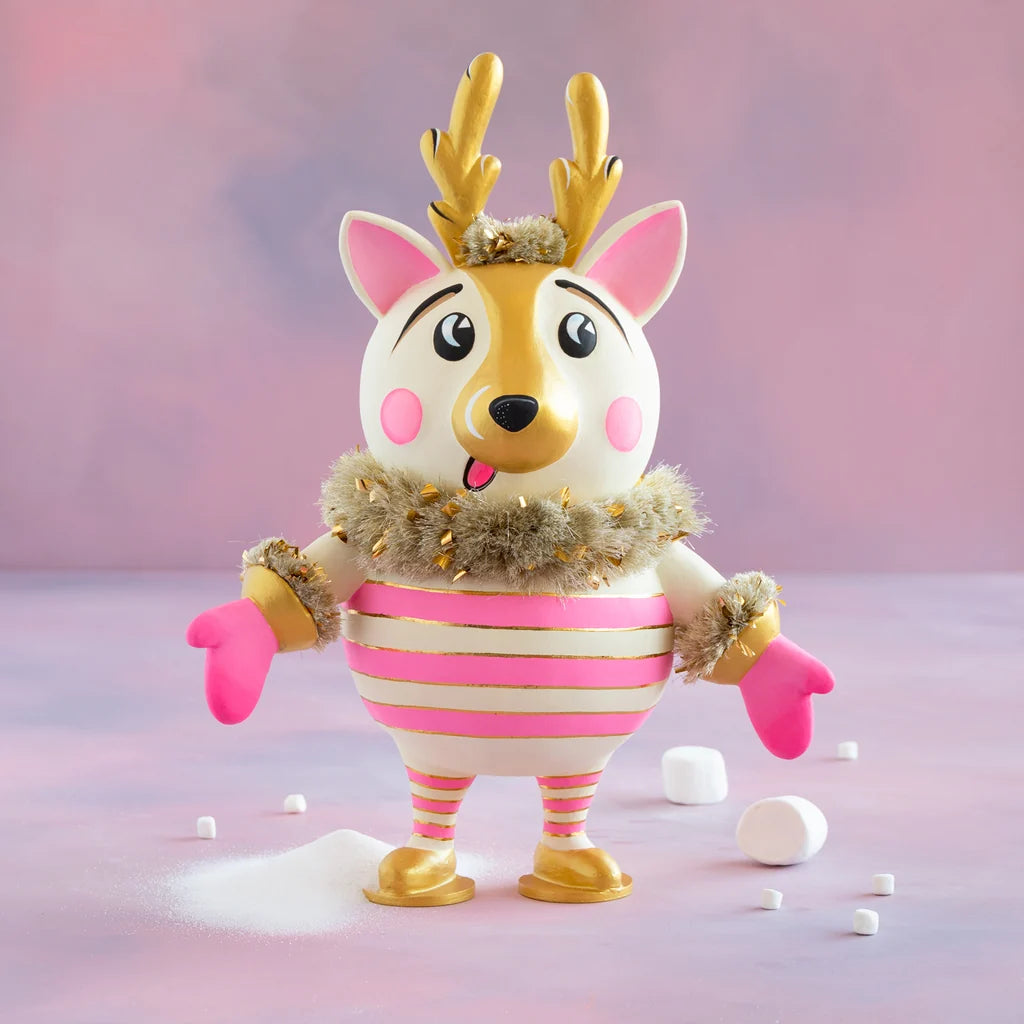 Cartoon-like La Renne reindeer tabletop display with a white and gold face, pink cheeks, gold antlers, pink and gold striped body, and fluffy gold tinsel accents against a soft pink snowy backdrop.