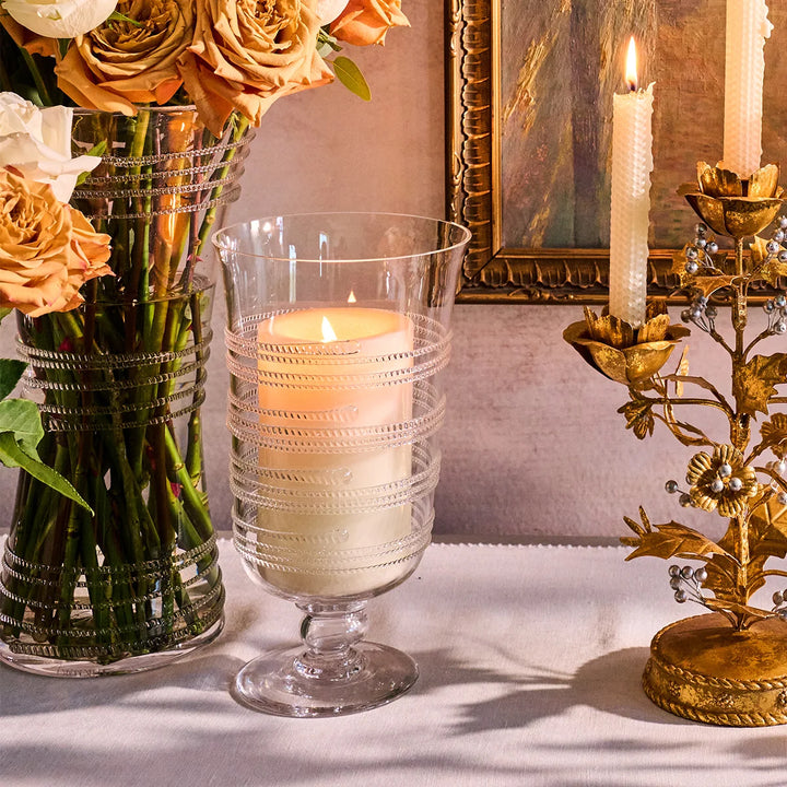 Clear glass hurricane candle holder with pillar candle styled beside flowers and gold floral candlesticks