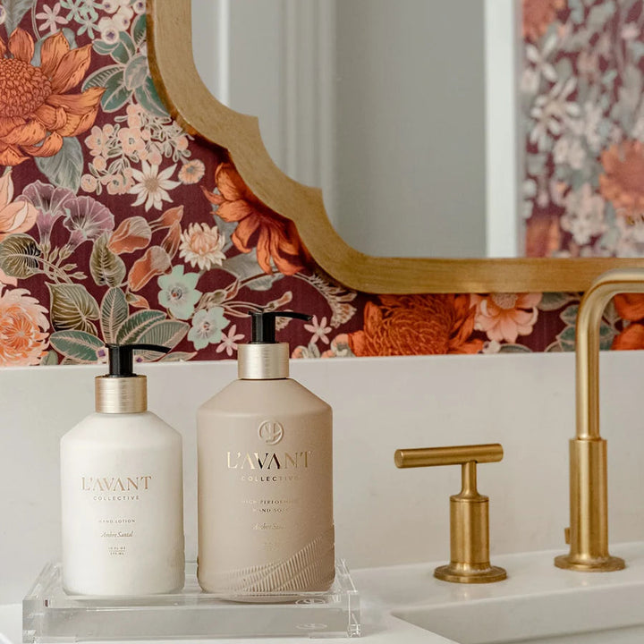 Clear lucite vanity tray displaying hand soap and hand lotion beside a bathroom sink with brass fixtures and floral wallpaper.