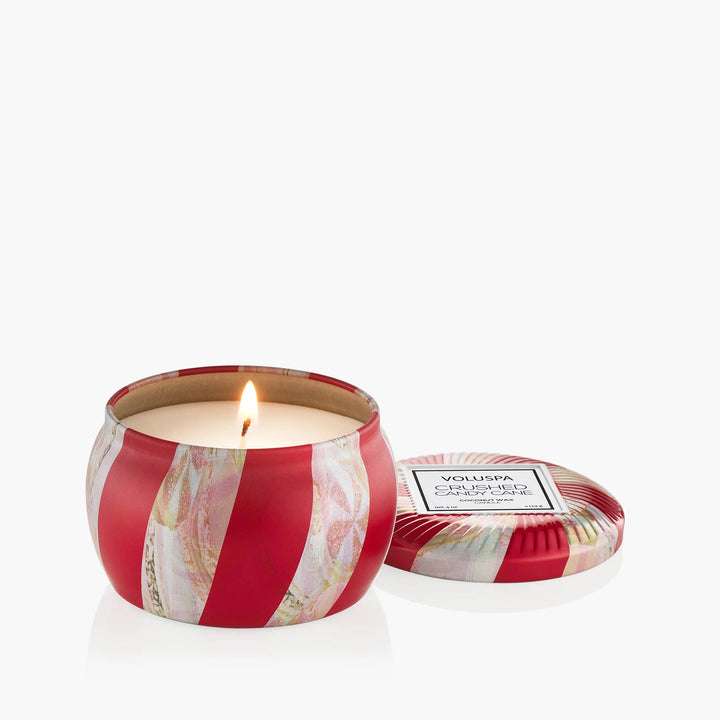 Crushed Candy Cane Mini Tin Candle by Voluspa lit beside its red and white peppermint-inspired lid