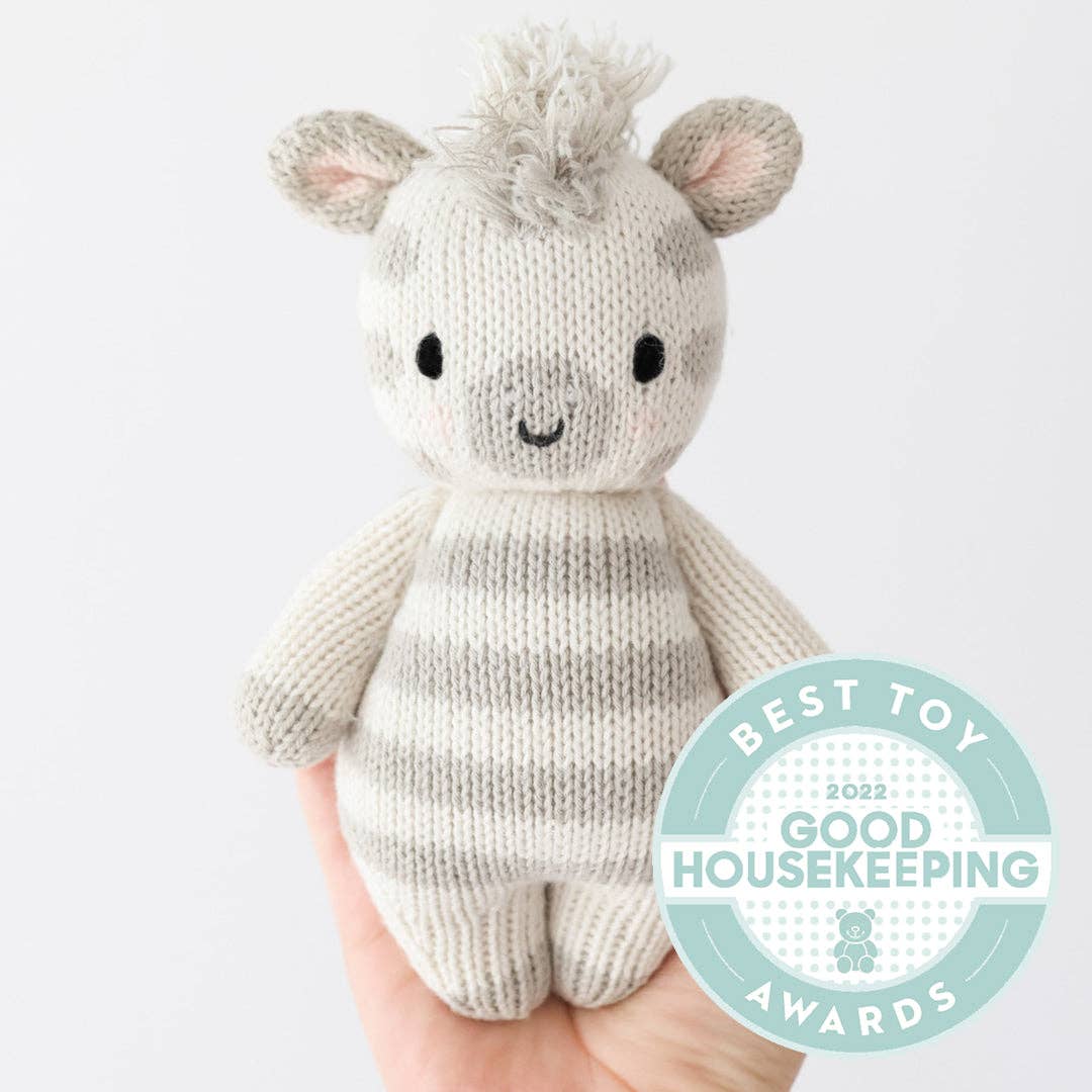 Cuddle+Kind Baby Zebra plush held upright by a hand, with a Best Toy housekeeping award in the lower right.