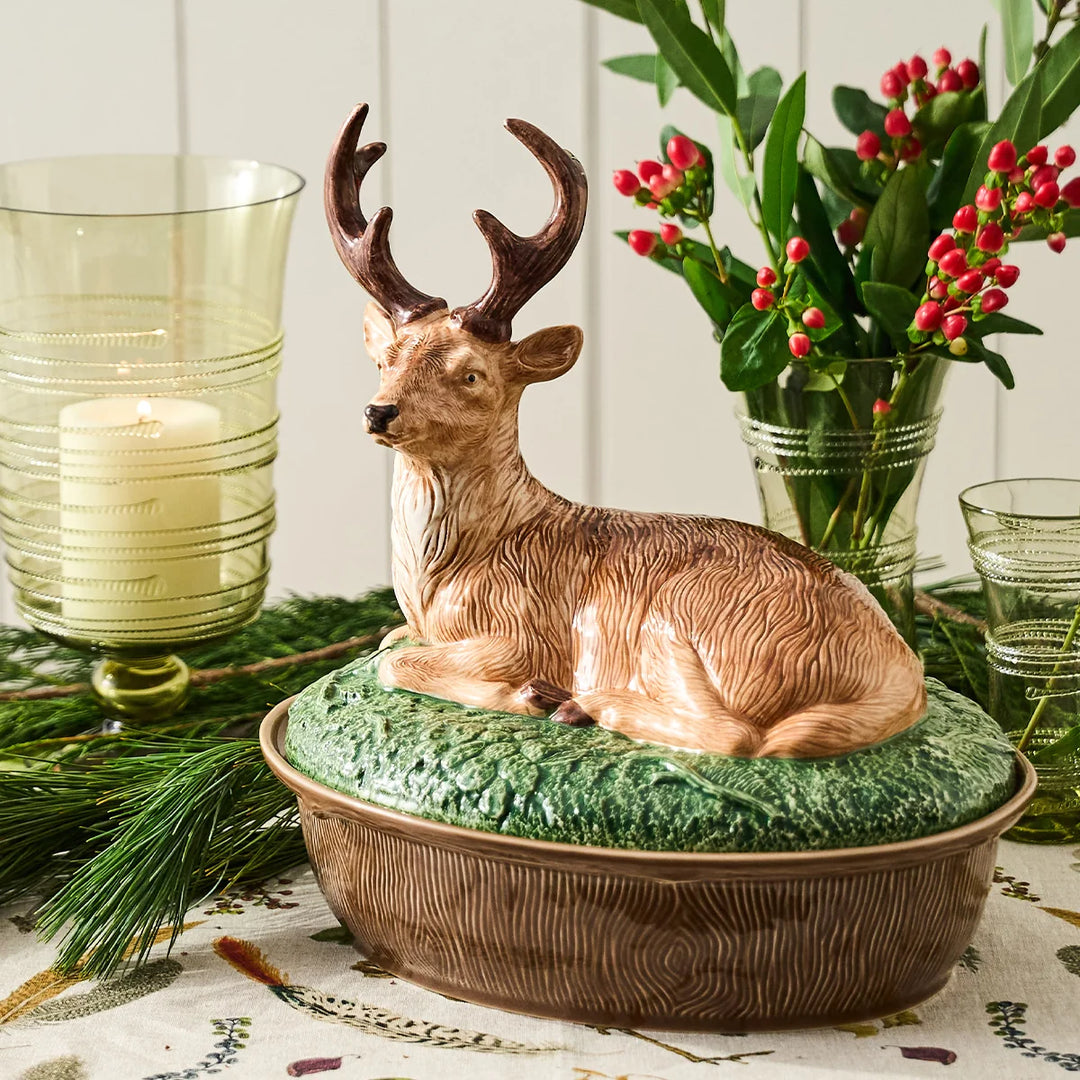 Ceramic deer covered serving dish displayed on a festive holiday table with evergreen branches, red berries and green glassware.