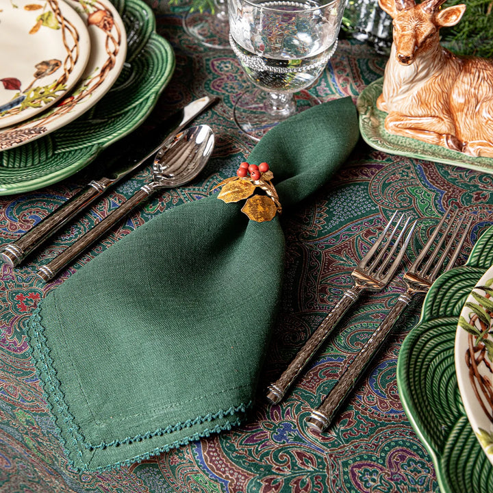 Forest green linen napkin with a gold holly and red berry napkin ring on a festive woodland holiday tablescape.