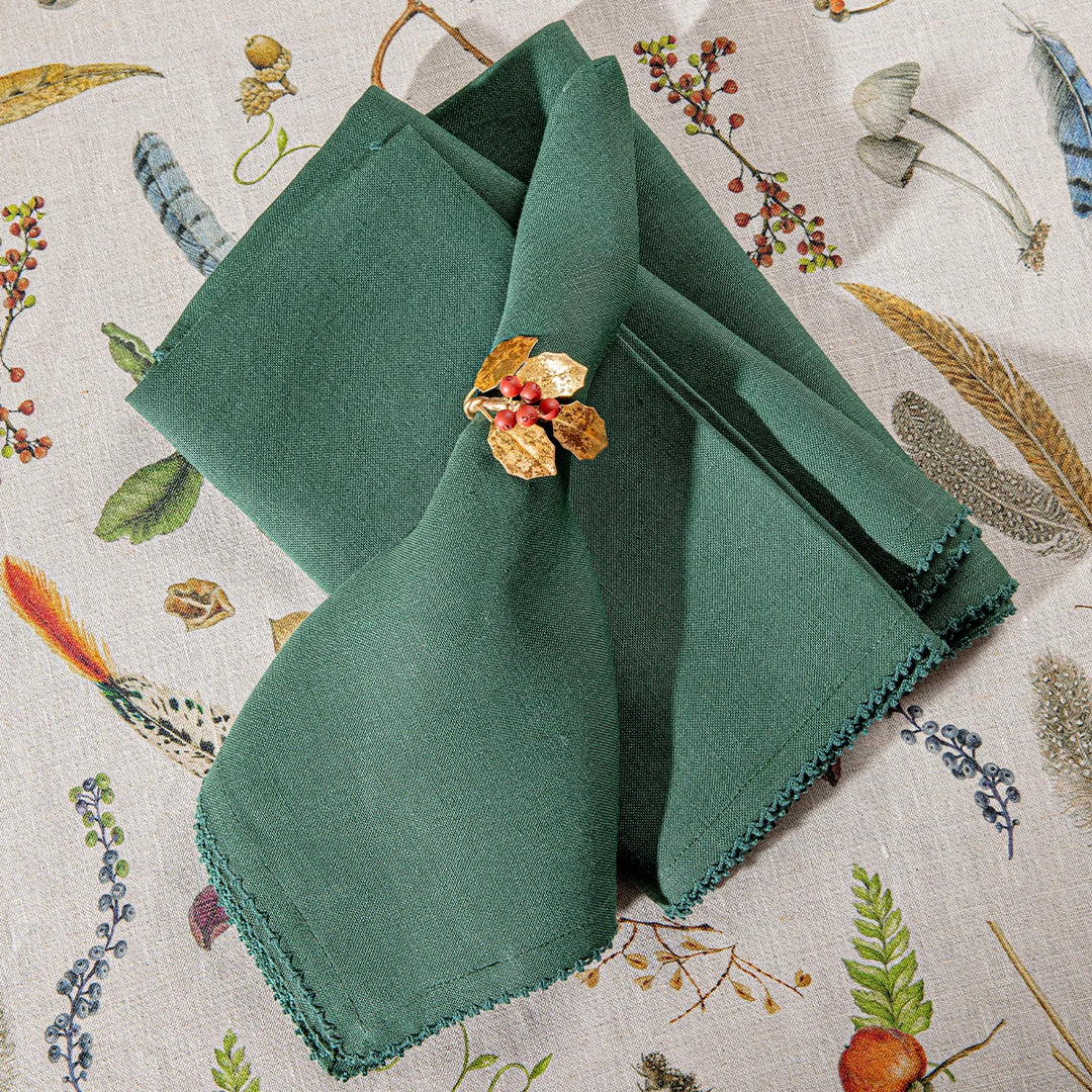 Forest green linen napkins styled with a gold holly and red berry napkin ring over a woodland patterned tablecloth.