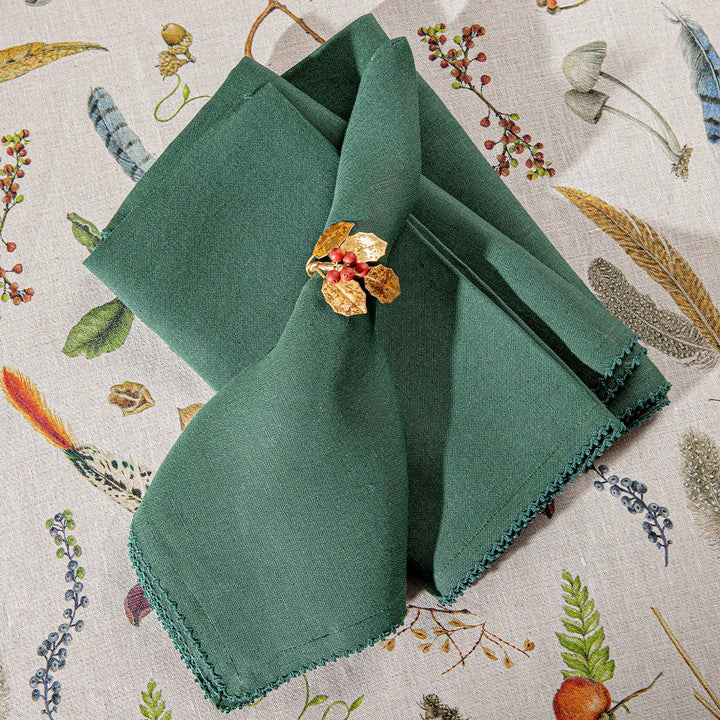 Forest green linen napkins styled with a gold holly and red berry napkin ring over a woodland patterned tablecloth.
