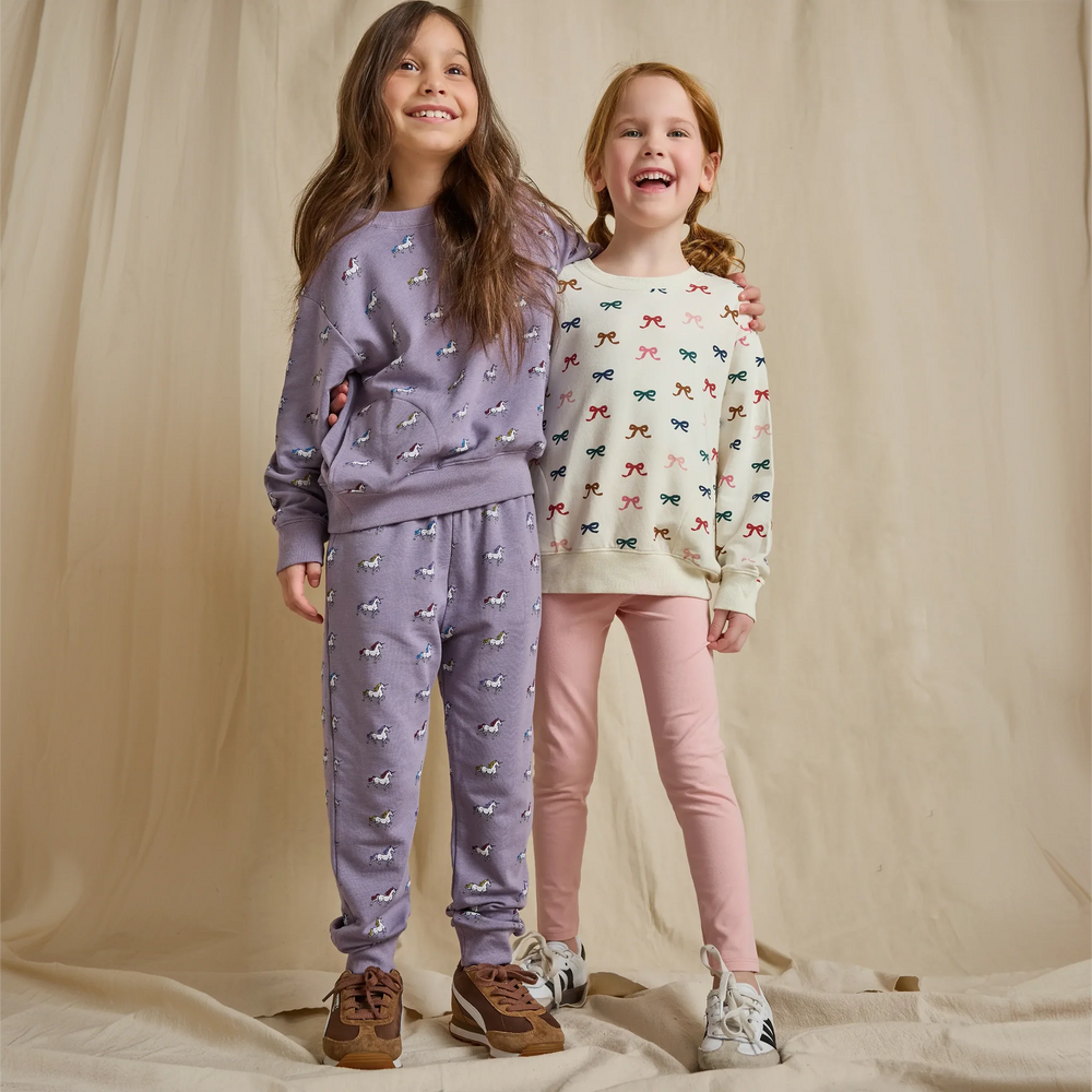 Girl wearing a cream multicolor bow sweatshirt with pink leggings alongside a girl in a purple unicorn outfit
