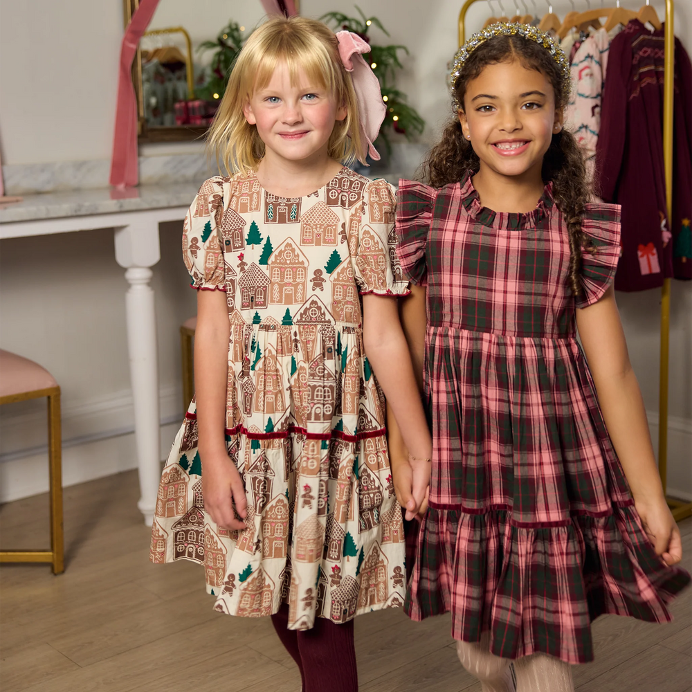 Young girl modeling the Pink Chicken Girls Eliza Dress in Gingerbread Houses with burgundy tights and a pink hair bow beside another girl in a plaid holiday dress.