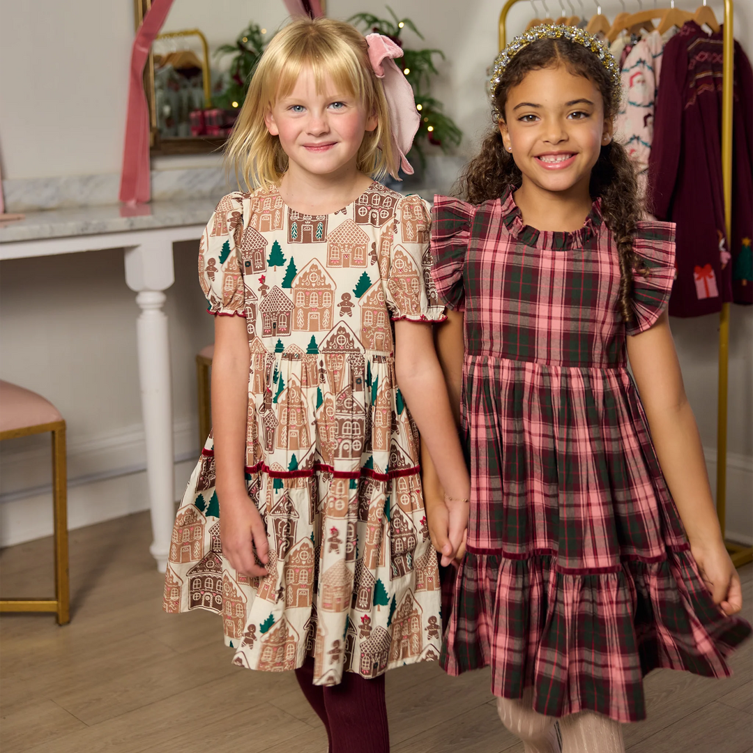 Young girl modeling the Pink Chicken Girls Eliza Dress in Gingerbread Houses with burgundy tights and a pink hair bow beside another girl in a plaid holiday dress.