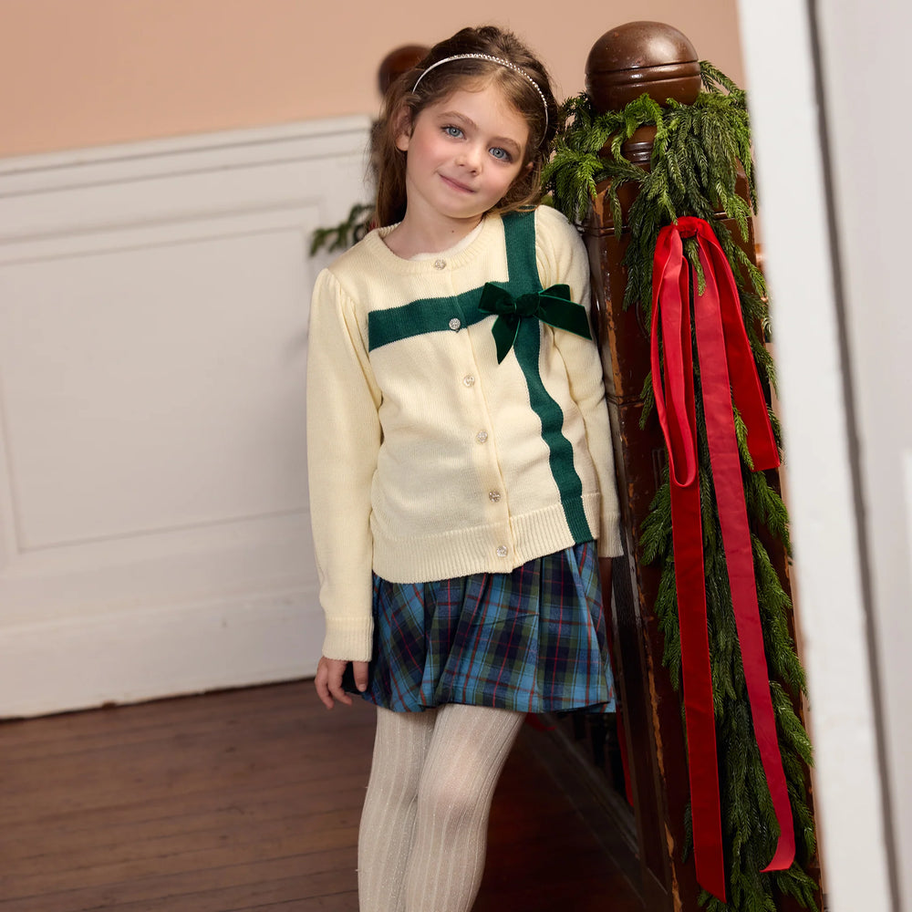 Girl wearing the Blue Tartan Gianna Skirt with a cream cardigan featuring green ribbon detailing for a festive holiday outfit