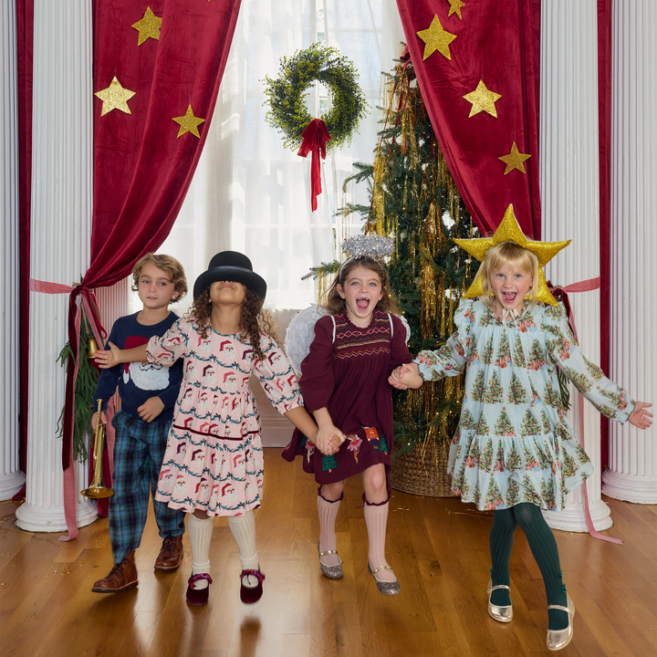 Girl wearing the Pink Chicken Girls Harper Dress in Trees and Presents with other children dressed in festive holiday outfits beside a Christmas tree.