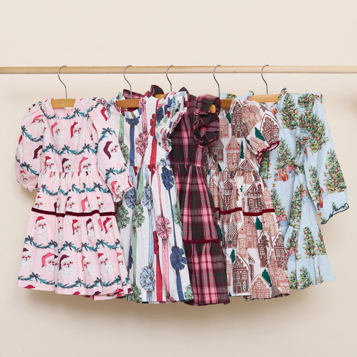 Pink Chicken holiday dress collection displayed on wooden hangers, including the Girls Harper Dress in Trees and Presents alongside festive Christmas dresses.