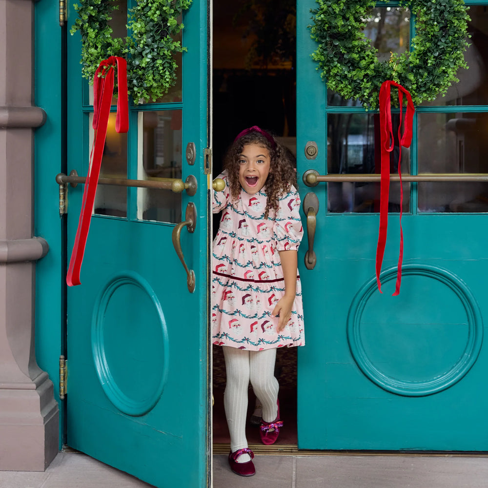 Girl wearing the Pink Chicken Maribelle Dress in Santa Garland while standing between festive teal doors decorated with Christmas wreaths.