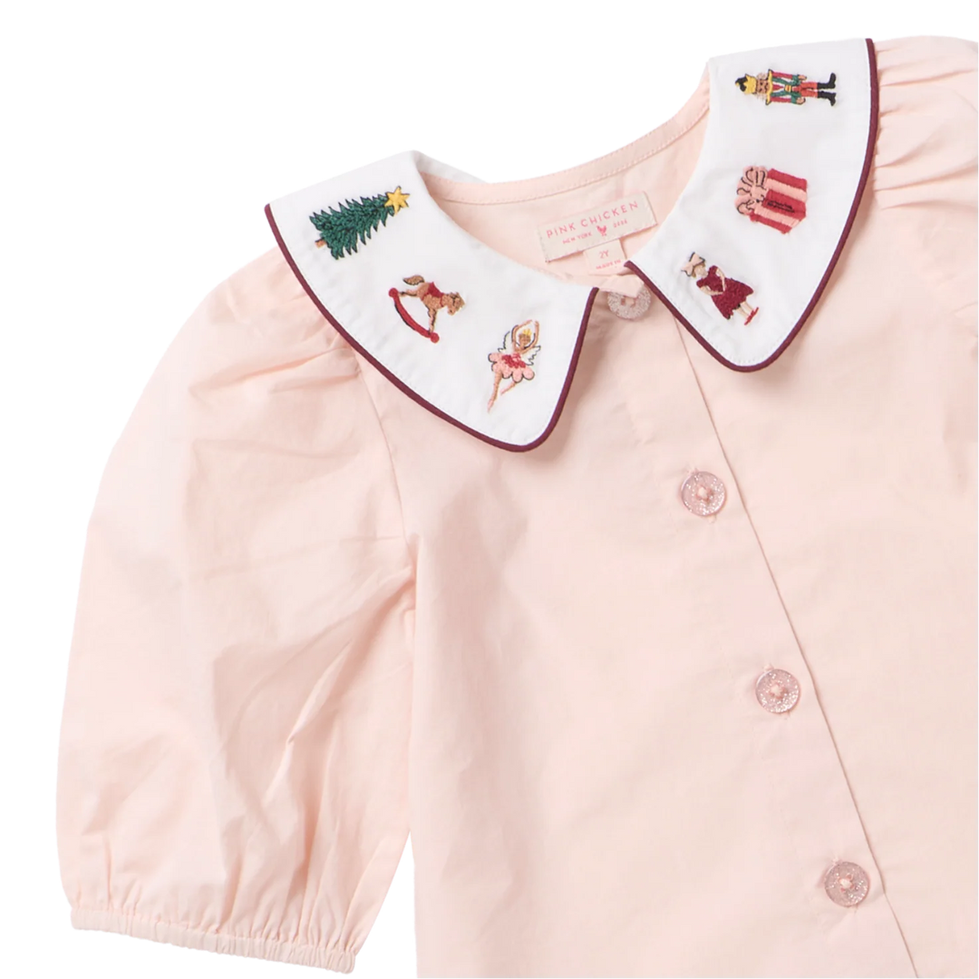 Close-up of the Girls Noelle Top featuring embroidered Christmas trees, a rocking horse, ballerina, nutcracker, and presents on a white collar with burgundy piping.