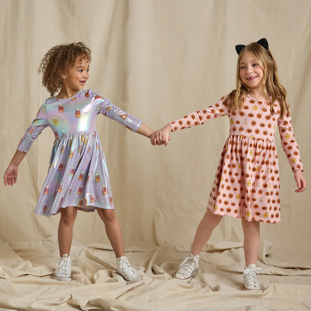 Girl modeling a pink pumpkin Halloween dress with long sleeves and jack-o'-lantern print alongside a lavender candy corn dress.