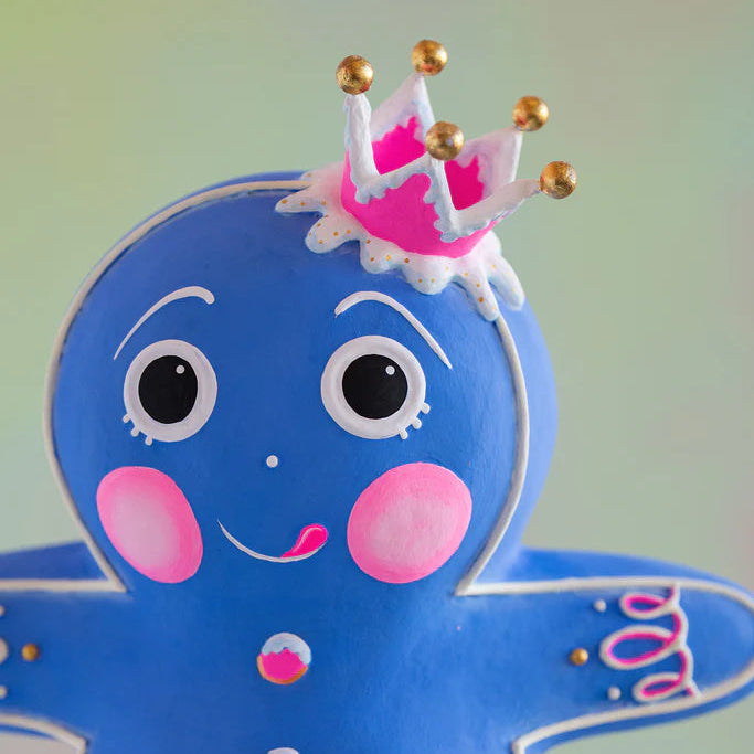 Close-up of the Glitterville Blue Dough gingerbread figure featuring a bright blue face, expressive white icing eyes and brows, pink cheeks, a playful smile, and a whimsical pink-and-white crown with gold ball accents.
