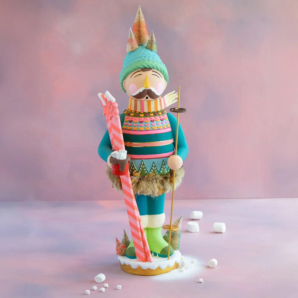 Glitterville Bronson display nutcracker dressed for winter, holding pink and orange skis and a ski pole while standing in the snow, with a colorful knitted hat, striped scarf, and festive alpine-inspired details.
