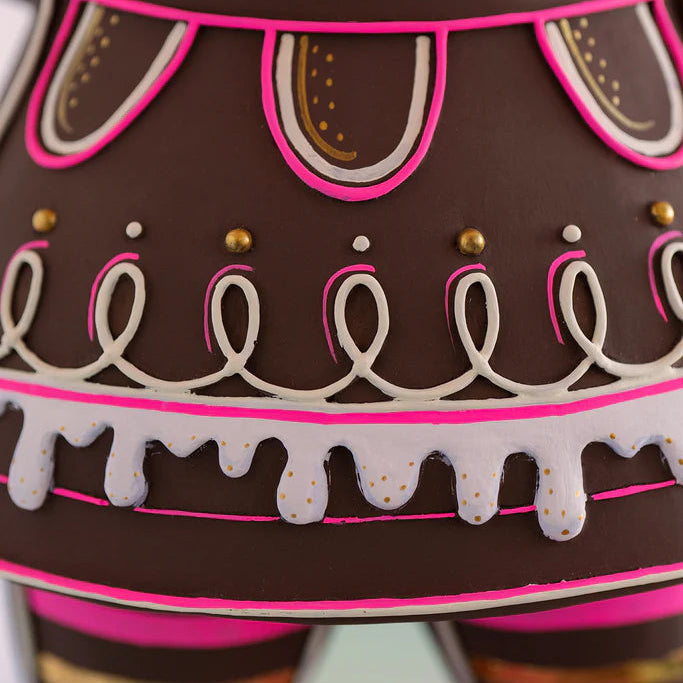 Glitterville Dippsy gingerbread figure dress close-up showcasing intricate pink, white, and gold icing details, including looping piped icing, decorative dots, scalloped icing trim, and bright pink accents on the brown gingerbread design.