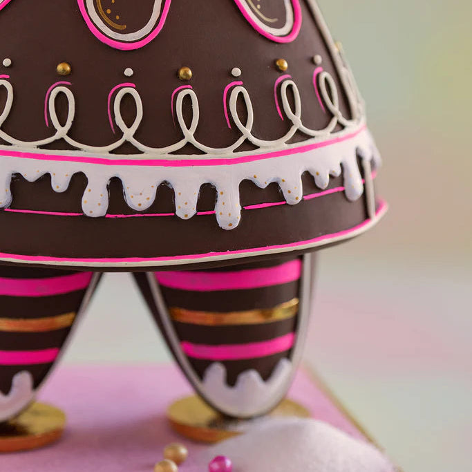 Glitterville Dippsy gingerbread figure close-up highlighting the detailed pink, white, and gold icing on her dress skirt and striped legs, including looping piped icing, scalloped white icing trim, gold decorative dots, bright pink accents, and gold shoes.
