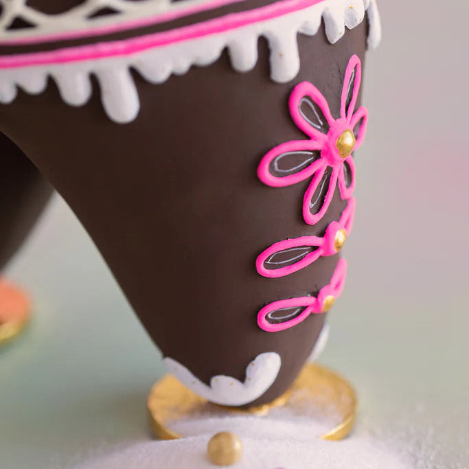 Glitterville Dough gingerbread figure leg close-up showcasing intricate pink, white, and gold icing details, including flower-shaped pink piping with gold centers, white icing trim, and gold shoe accents on the brown gingerbread design.