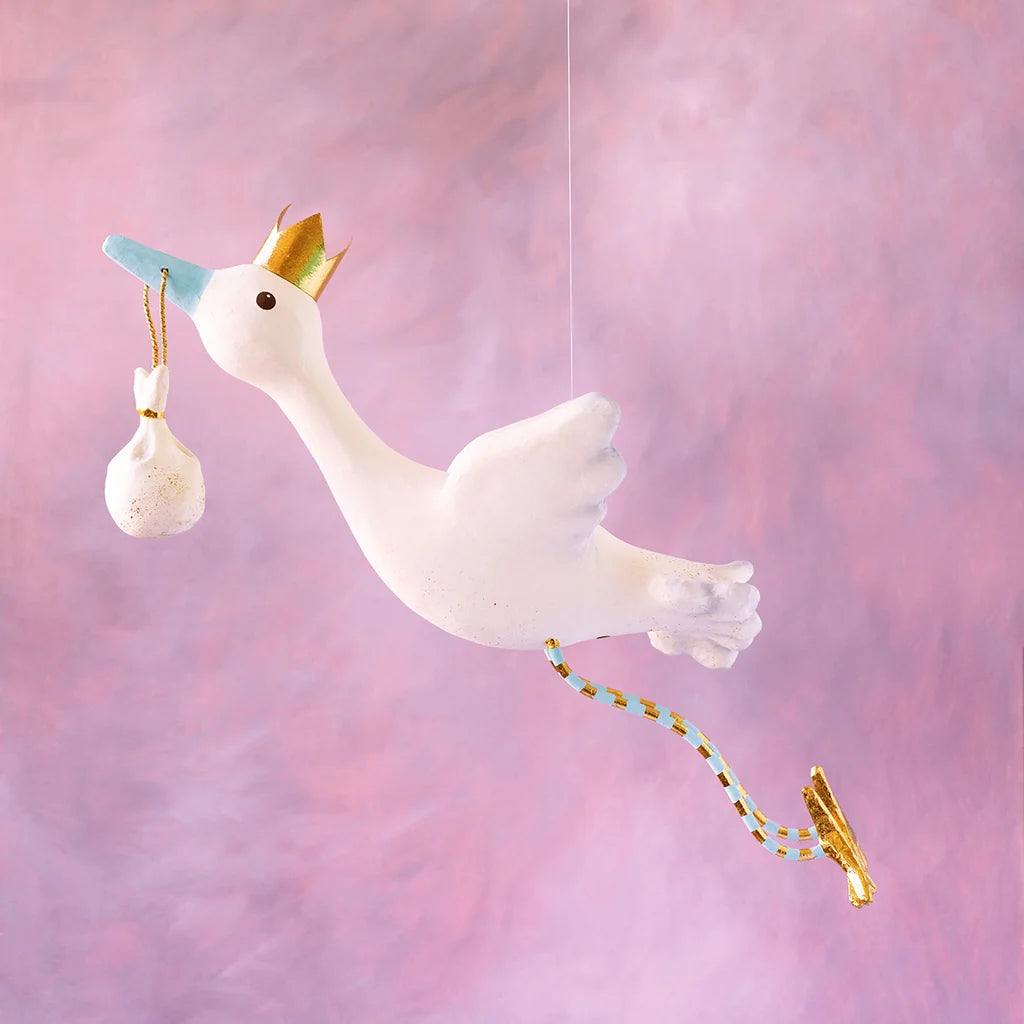 Glitterville Flying Stork Display in white and blue, featuring a gold crown, dangling baby bundle, and gold accents against a pink background.