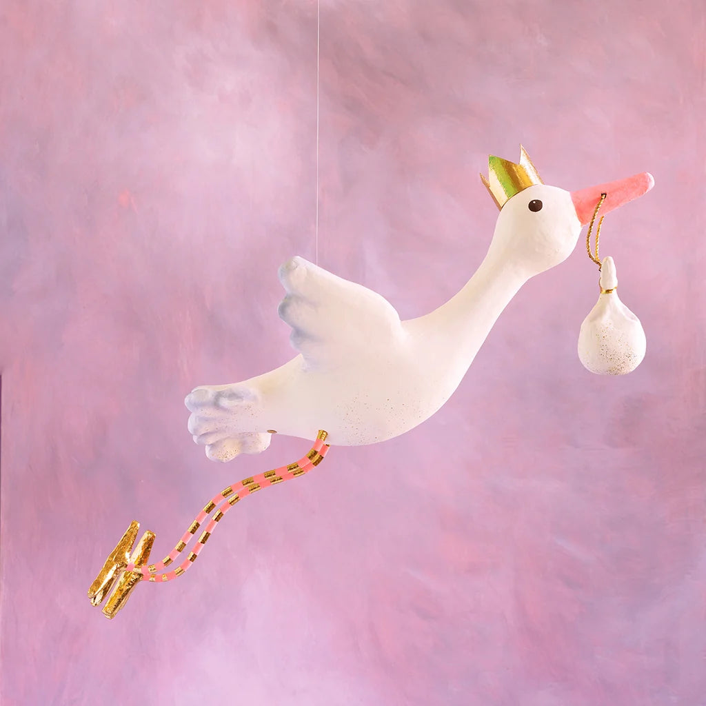 Glitterville Flying Stork Display in white and blue, featuring a gold crown, dangling baby bundle, and gold accents against a pink background.