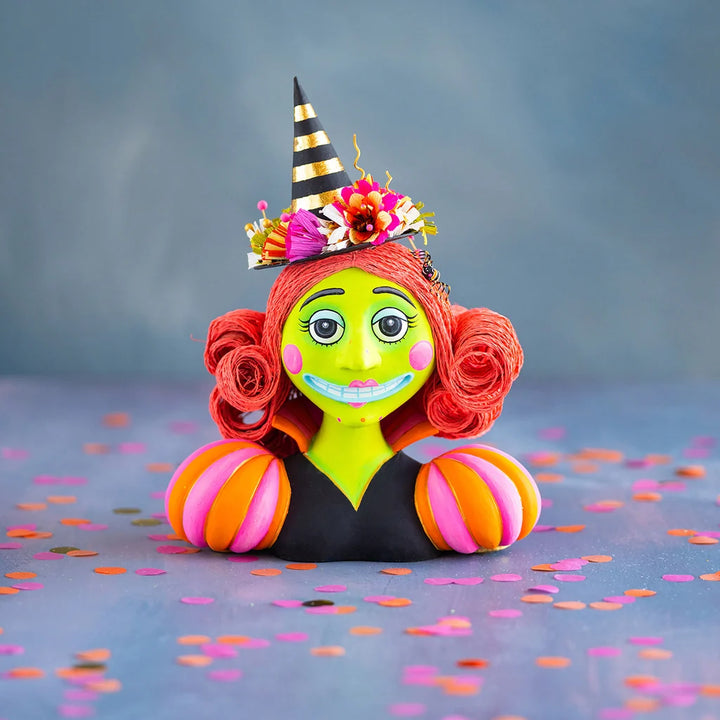 Glitterville Itchy Witchy Bust tabletop Halloween decoration featuring a whimsical lime-green witch with bright red curled hair, a black and gold striped pointed hat decorated with colorful flowers, pink and orange striped sleeves, and a playful spooky smile.