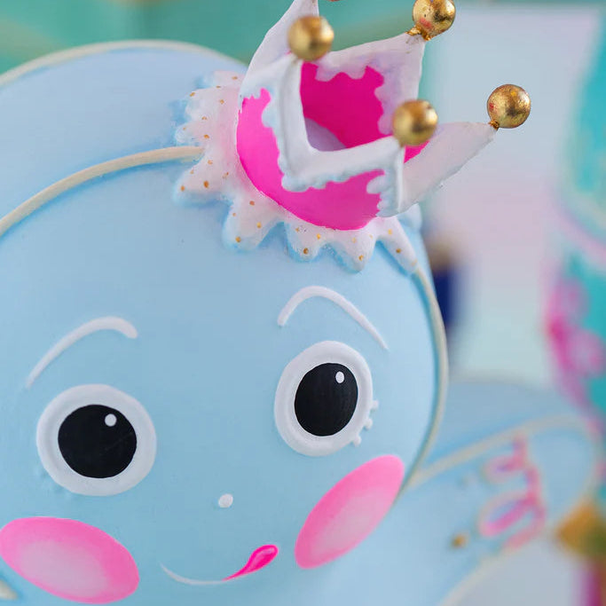 Close-up of the Glitterville Light Blue Dough gingerbread figure featuring a whimsical pink and white icing crown topped with gold accents, detailed white icing around the crown, expressive piped eyes and brows, rosy pink cheeks, a sweet pink smile, and delicate decorative icing on the arm. This detailed view highlights the intricate handcrafted-style icing and pastel holiday design of the Glitterville gingerbread Christmas collectible.