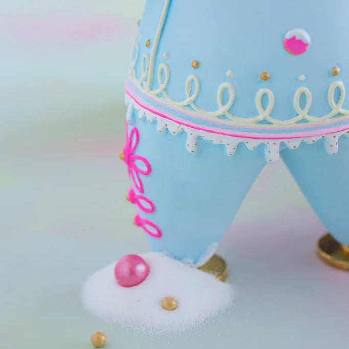 Close-up of the Glitterville Light Blue Dough gingerbread figure highlighting the intricate icing details on the legs, including bright pink piped loop designs with gold accents, white dripping icing trim, pink and white candy buttons, gold details, and metallic gold shoes. This detailed product photo showcases the whimsical handcrafted-style decoration and pastel Christmas design of the Glitterville gingerbread collectible.