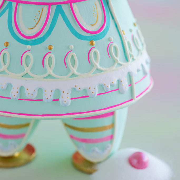 Close-up of the Glitterville Mint Dipsy gingerbread figure featuring the detailed pastel dress skirt, white and pink looped icing, dripping white icing trim with gold sprinkles, striped legs, and gold shoes.